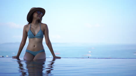 Pretty-young-Asian-woman-in-a-bikini-leans-against-the-edge-of-a-resort-pool