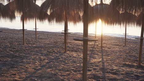 Slow-pan-right-at-Marbella-beach-during-sunrise-with-beautiful-tiki-straw-sun-umbrellas-on-beach,-bright-sunrise-giving-warm-look-and-feel-to-4k-footage