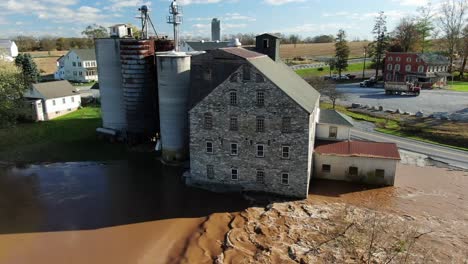 Aerial-of-flooded-grain-mill,-silos,-elevators-beside-muddy-river-on-sunny-day