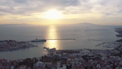 Aerial-drone-Flight-over-downtown-mitilini-revealing-sunrise-at-sea-with-ferry-to-athens,-lesvos,-greece