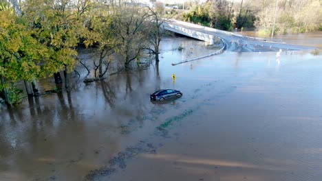 Flooded-blue-car-stranded-in-swollen-river-covering-road-and-bridge,-aerial-push-in-shot
