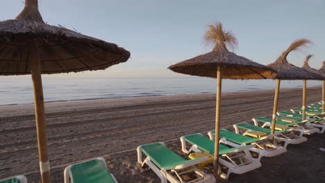 Slow-panning-left-on-Marbella-beach-at-sunrise-with-sunbeds-and-straw-umbrellas,-tranquil-small-waves-on-an-empty-beach,-dream-luxury-travel-inspiration