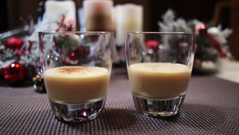 Two-glasses-of-Christmas-eggnog-with-ground-cinnamon-being-added-garnished-with-Christmas-decorations
