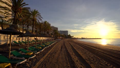 POV-walking-down-Marbella-beach-at-sunrise-with-gimbal,-left-side-with-sunbeds-and-parasols-with-tropical-palm-trees,-right-side-Mediterranean-sea,-luxury-travel-inspiration