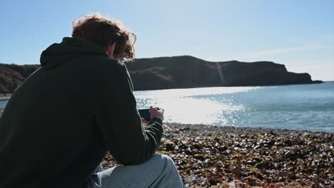 A-young-watches-his-phone-on-lulworth-cave-beach,-morning-landscape