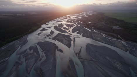 aerial-flying-backwards-over-the-braided-Waimakariri-river-with-the-sun-setting-providing-golden-light-on-the-channels,-Christchurch,-New-Zealand
