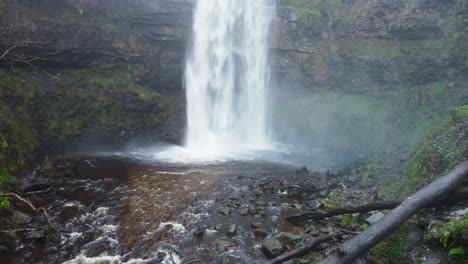 Luftaufnahme-Eines-Großen-Wasserfalls-Mit-Gehackten-Baumstämmen-Und-Einer-Höhle-Hinter-Dem-Wasser-In-Wales,-Großbritannien,-4k