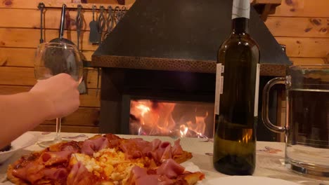 Hand-Reaching-Out-To-Pick-Up-Wine-Glass-Beside-Rustic-Pizza-With-Fireplace-In-Background