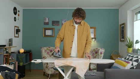 Modern-Caucasian-man-ironing-clothes-in-stylish-living-room-doing-housekeeping-chores