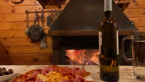 Hand-Reaching-Out-To-Pick-Up-Wine-Glass-From-Table-Beside-Rustic-Pizza-With-Fireplace-In-Background-In-Wood-Cabin