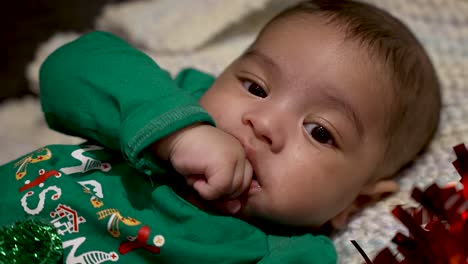 Adorable-Cute-Indian-Baby-Wearing-Green-Festive-Christmas-Top-Sucking-Left-Hand-As-He-Lays-on-Blanket