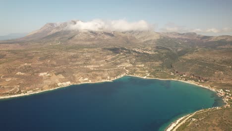 Eine-Drohne-Fliegt-Langsam-In-Großer-Höhe-Neben-Dem-Blauen-Meer,-Während-Die-Berge-An-Einem-Heißen-Sommertag-In-Griechenland-Mit-Wolken-Bedeckt-Sind