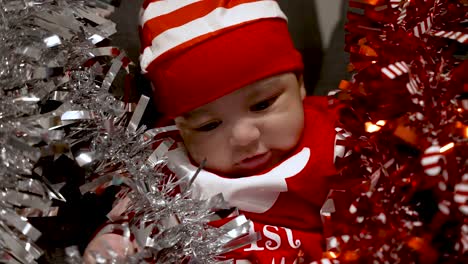 Adorable-2-Month-Old-Baby-Boy-Wearing-Red-Christmas-Outfit-And-Hat-And-Being-Playful-With-Red-And-Silver-Tinsel-Surrounded-Him-On-Sofa