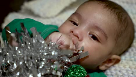 Adorable-2-Month-Bangladeshi-Baby-Boy-Lying-On-Blanket-Sucking-Hand-Whilst-Grabbing-Silver-Tinsel