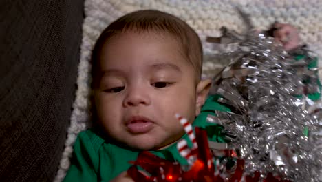 Overhead-View-Of-Adorable-Cute-Indian-Baby-Wearing-Green-Festive-Christmas-Top-Pulling-Silver-And-Red-Tinsel-Over-Him