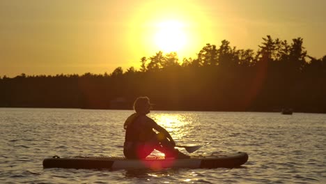 pretty-girl-sitting-on-paddle-board-strong-sunset-with-lens-flare-slomo-epic