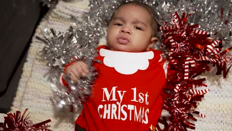 Overhead-View-Of-Cute-Two-Month-Old-Baby-Wearing-Red-My-1st-Christmas-Outfit-Laid-On-Blanket-Pulling-And-Grabbing-Red,-Silver-Tinsel
