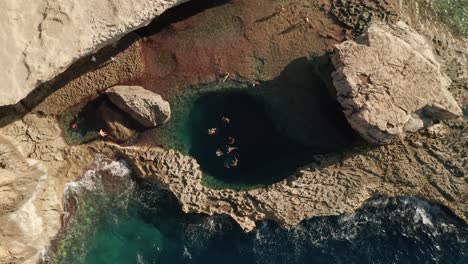 Aus-Der-Luft-Herauszoomen-Ansicht-Von-Menschen,-Die-In-Einem-Unberührten-Blauen-Meer-Im-Berühmten-Blauen-Loch-In-Dwerja-Auf-Der-Insel-Gozo,-Malta,-Schwimmen