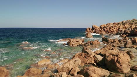 Aerial-dolly-shot-of-a-seashore-with-beautiful-turquoise-waters-of-the-Mediterranean-Sea-splashing-on-the-red-coloured-rocks