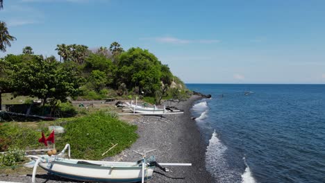 Canoas-De-Pesca-Tradicionales-Balinesas-Esparcidas-En-Una-Playa-De-Guijarros-Volcánicos-Negros-Cerca-De-Un-Pequeño-Pueblo