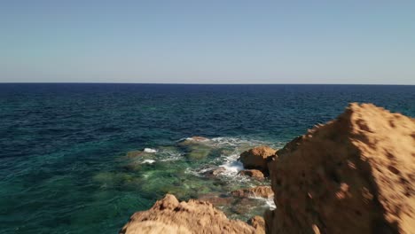 Aerial-dolly-shot-of-a-seashore-with-beautiful-turquoise-waters-of-the-Mediterranean-Sea-splashing-on-the-red-coloured-rocks