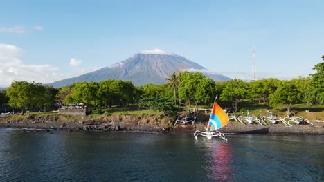 Die-Farbenfrohen-Segel-Der-Traditionellen-Balinesischen-Fischerboote-Segeln-Zu-Den-Küstenstränden,-Die-Vom-Hoch-Aufragenden-Vulkan-Mount-Agung-In-Den-Schatten-Gestellt-Werden