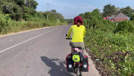 Vista-Trasera-De-Mujeres-En-Bicicleta-En-La-Calle-De-Laos-Con-árboles-En-Ambos-Lados-De-La-Calle