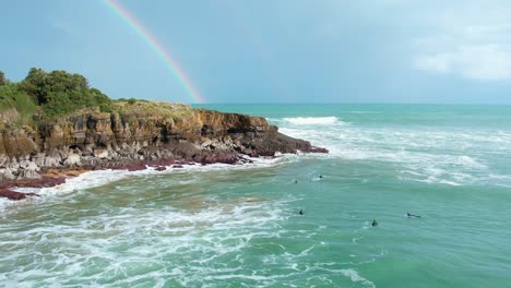 Drone-shot-of-the-waves-crashing-the-cliffs-on-the-beach,-with-a-rainbow-in-the-background