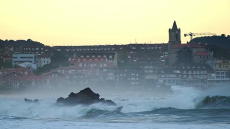 Big-waves-breaking-over-the-rocks-with-the-city-buildings-in-the-background-in-the-evening