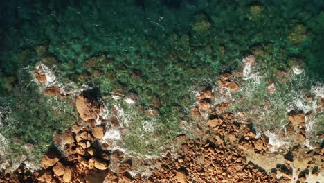 Aerial-zoom-in-view-of-clear-turquoise-sea-and-waves-crushing-on-red-colored-rocks-on-the-beach-near-Rampla-Beach-on-the-island-of-Gozo,-Malta