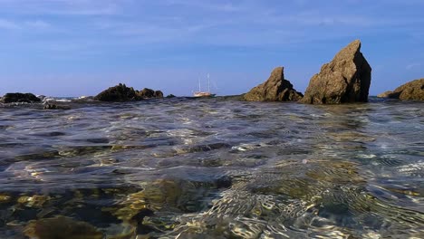 Meerwasseroberflächenperspektive-Des-Segelschiffs,-Das-Am-Horizont-Festgemacht-Ist,-Mit-Segeln,-Die-Zwischen-Felsen-Gesenkt-Werden