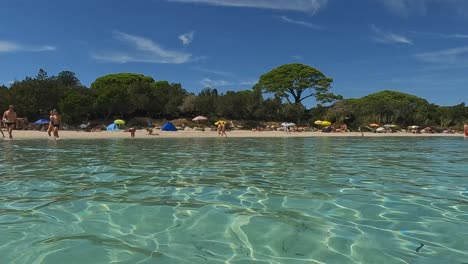Low-angle-sea-water-surface-pov-of-Santa-Giulia-beautiful-beach-in-Corsica-island-with-people-on-shore-on-summer-day,-France