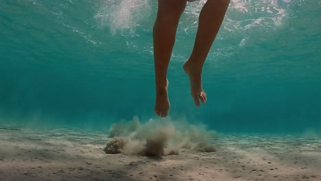 Underwater-scene-of-little-girl-jumps-on-seabed-raising-clouds-of-sand-in-transparent-water