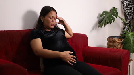 Mujer-Embarazada-Asiática-Con-Dolor-De-Cabeza-Mientras-Acaricia-El-Estómago-En-Casa