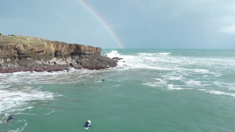 Unrecognizible-people-looking-at-waves-crashing-at-the-cliffs,-with-a-rainbow-in-the-background
