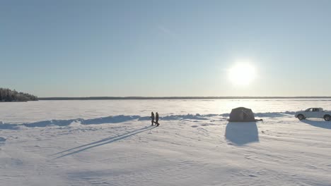 2-women-walking-away-from-tent-ice-fishing-on-a-frozen-lake,-tundra,-drone