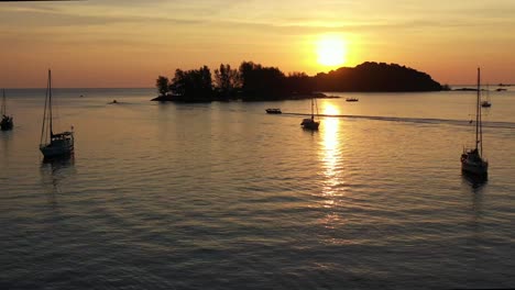 Sailing-sunset-landscape-view,-aerial-forward-flying-capturing-glowing-sun-setting-behind-seratos-island-and-paradise-101,-reflection-on-calm-sea-ocean,-tropical-paradise-at-Langkawi,-Kedah,-Malaysia
