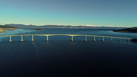 A-long-box-girder-bridge-floating-over-a-fjord-in-Norway-with-a-lone-car-driving-over-it---tilt-down-aerial-view