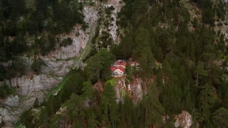 a-drone-pans-slowly-around-a-stone-cabin-surrounded-with-trees-on-a-cliff-under-the-peak-of-a-mountain