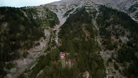 a-drone-flies-around-a-small-stone-cabin-that-is-surrounded-with-trees-on-a-cliff-under-the-peak-of-a-mountain