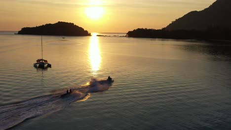 Sunset-silhouette-of-tourists-on-banana-boat-ride,-sailing-across-the-sea-with-water-splashing,-glowing-sun-setting-below-the-horizon,-shimmering-golden-reflection-on-ocean-surface,-Langkawi-island