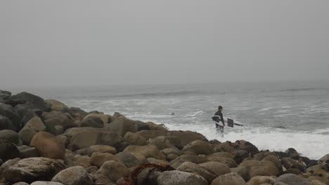 Surfer-standing-on-rocky-ocean-shore-as-he-gets-ready-to-paddle-out-to-sea