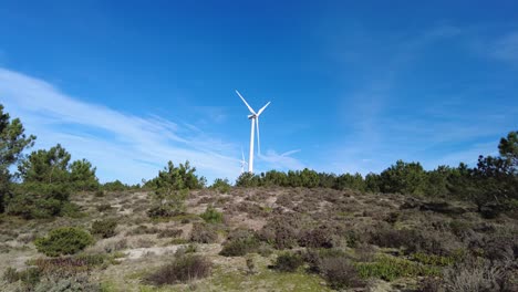 Two-wind-turbines-spinning-with-green-landscape-in-foreground-and-blue-sky-above