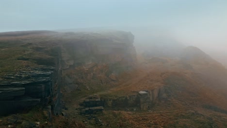 Aerial-drone-view-of-an-clouds-moving-over-Standedge-Edge-in-the-hills,-Pennines-on-a-foggy-morning,-golden-hills-and-beautiful-rocky-cliffs-and-moorlands
