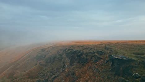 Aerial-drone-view-of-morning-mists-moving-slowly-over-the-rugged-Pennine-hill-tops,-on-a-foggy-morning,-golden-hills-and-beautiful-rocky-cliffs-and-moorlands