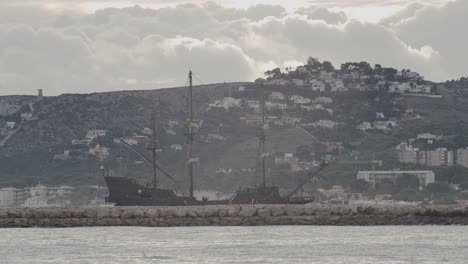 16th-Century-Galleon-Andalucia-replica-ship-arriving-at-port-in-a-beautiful-cloudy-day-at-sunrise-behind-a-breakwater-with-mountain-and-houses-in-the-background
