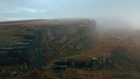 High-up-aerial-view-in-the-clouds,-mist-moving-in-over-the-cliff-tops,-foggy-golden-hills-and-beautiful-rocky-outcrop-and-moorlands