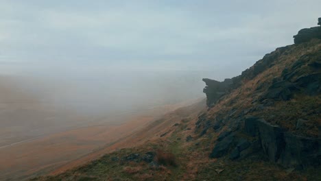 Aerial-drone-view-of-morning-mists-moving-slowly-over-the-rugged-Pennine-hills,-on-a-foggy-morning,-golden-hills-and-beautiful-rocky-cliffs-and-moorlands