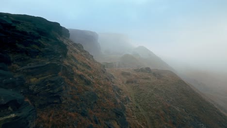Aerial-drone-view-of-morning-mists-moving-slowly-over-the-Pennine-hills,-on-a-foggy-morning,-golden-hills-and-beautiful-rocky-cliffs-and-moorlands