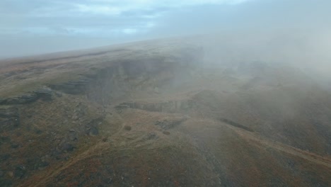 Aerial-drone-view-of-clouds-moving-slowly-over-the-Pennine-hills,-on-a-foggy-morning,-with-golden-hills-and-beautiful-rocky-outcrops-and-moorlands
